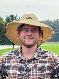 A man wearing a straw hat standing in a field with trees in the background.